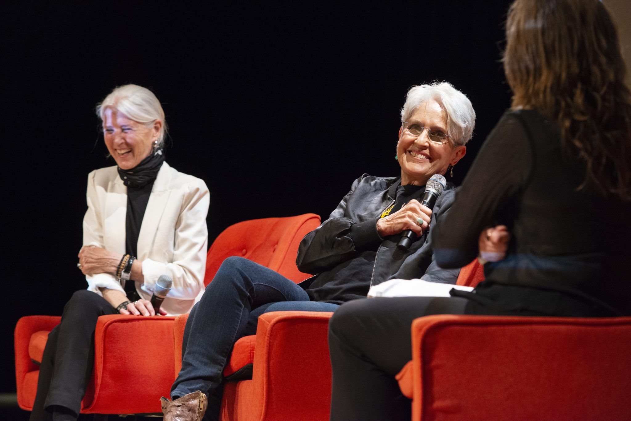 Joan Baez on stage at DC/DOX festival sitting in red chairs among interviewers and panelists laughing with microphone in hand