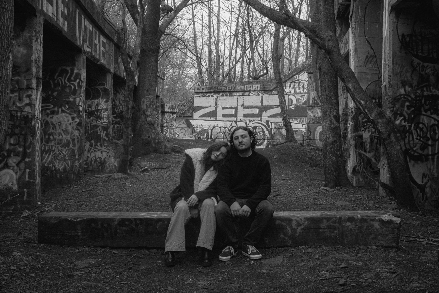 Black and white photo of GLOSSER sitting on a piece of wood surrounded by nature and graffiti as Fanning has her head on Sheehan's shoulder.