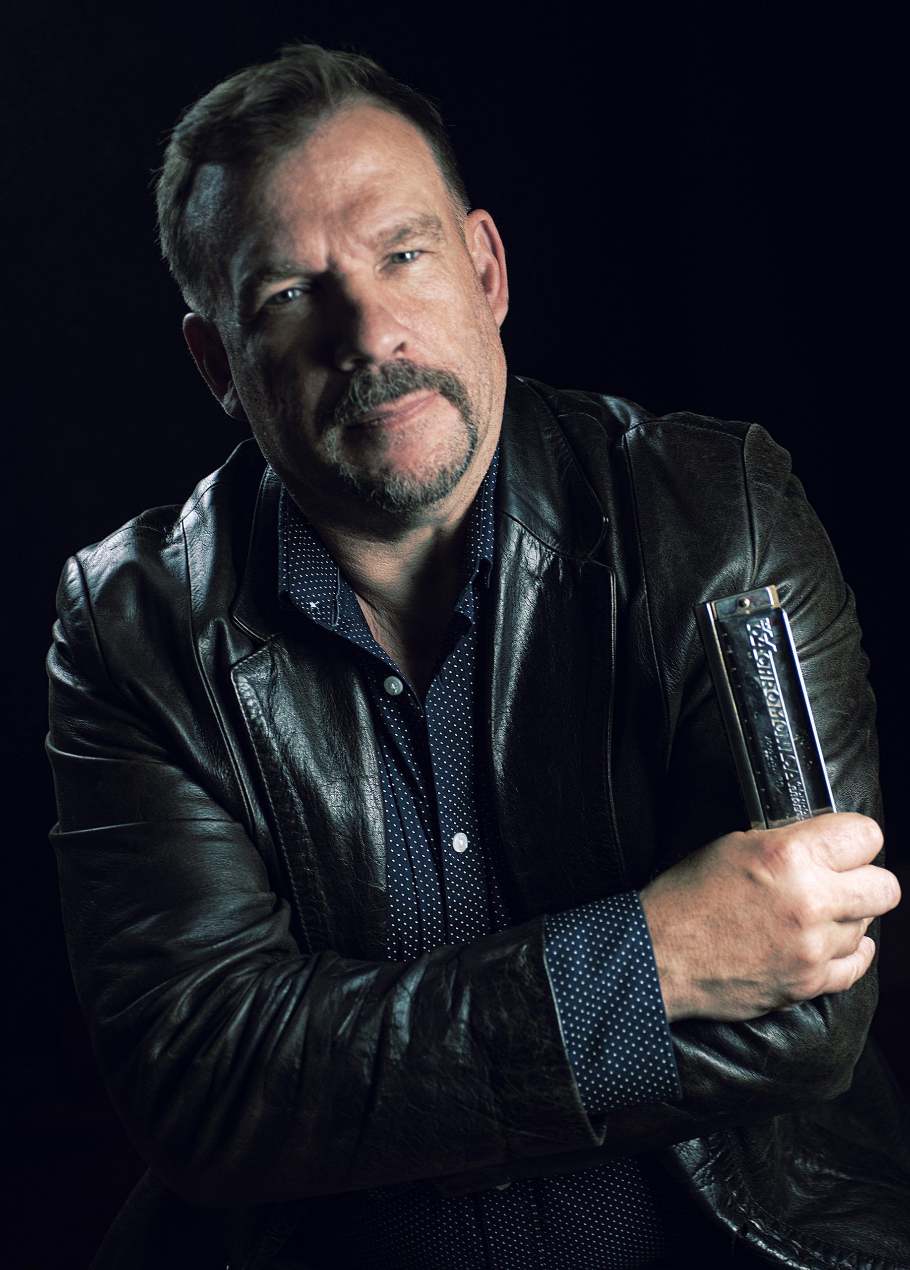 Chris O'Leary poses for profile photo in dramatic lighting while holding the harmonica.