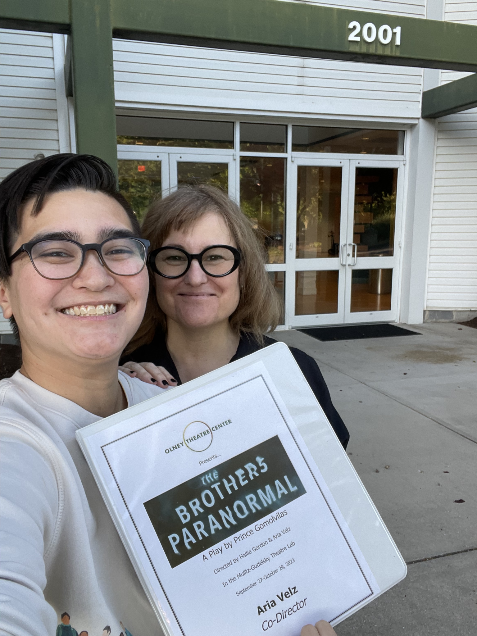 Aria Velz and Hallie Gordon holding a copy of 'The Brothers Paranormal' Script in front of Olney Theatre Center.