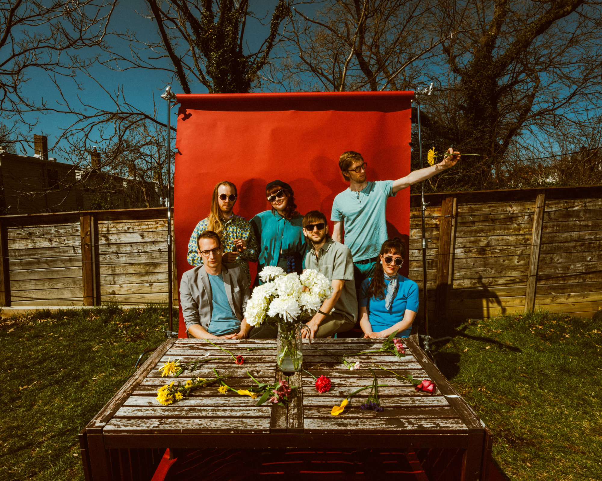 The North Country band sits together outside in front of a red photo backdrop. A wooden table in front of the band displays yellow and white flowers.