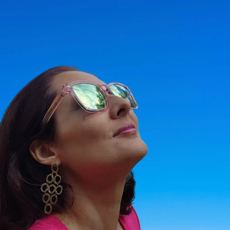 Velu O looks up to the sky in a pink shirt with sunglasses against a clear blue background.